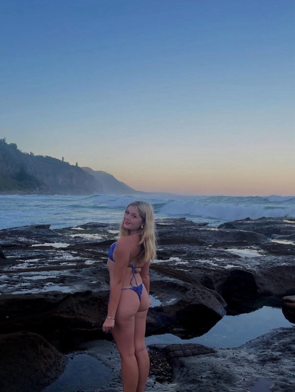 Woman in a blue bikini standing on rocky beach with ocean and sunset in the background twahli swim sustainable bikini 
